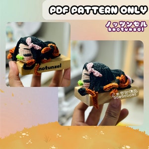 Może przedstawiać: Dzianinowa figurka postaci z anime Demon Slayer, leżąca na drewnianej podstawie. Figurka ma na sobie czarny i pomarańczowy strój z różowymi akcentami. Podstawa jest oznaczona jako "knotunzel".