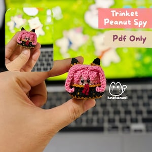 May include: A small crocheted figure with pink hair, black ears, and a black and gold outfit with a red accent. The figure features two black bead eyes. Text on the image reads Trinket Peanut Spy Pdf Only and includes a cat logo with the text knotunzel.