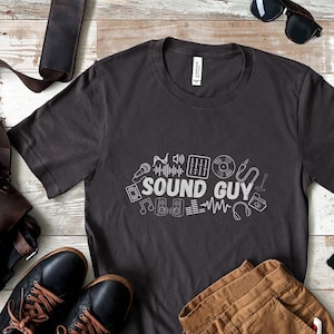 Puede incluir: Una camiseta gris oscuro con un diseño gráfico blanco de equipo de música y el texto "SOUND GUY".