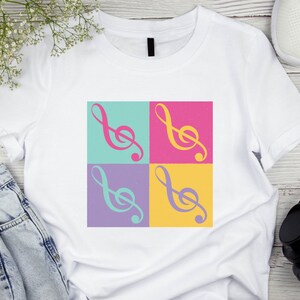 May include: White t-shirt featuring a colorful pop art design of a treble clef in four squares. The clef is in pink, yellow, and teal, set against a background of teal, pink, purple, and yellow squares. The shirt is paired with jeans and sneakers.