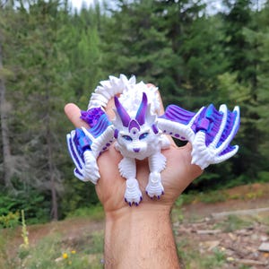 May include: A white articulated dragon figurine featuring vibrant purple and blue iridescent accents on its wings, ears, and tail. The creature has a feline-like face with blue eyes and a segmented body structure designed for flexible movement.