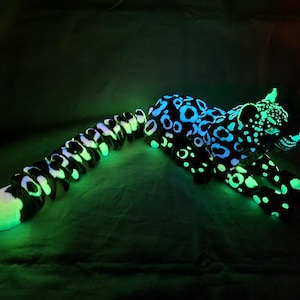 Könnte beinhalten: Ein Plüsch-Leopard-Spielzeug mit leuchtendem Neon-Design. Der Leopard hat einen schwarzen Körper mit blauen und grünen Flecken und einen segmentierten Schwanz mit abwechselnd schwarzen und weißen Abschnitten. Das Spielzeug leuchtet im Dunkeln.