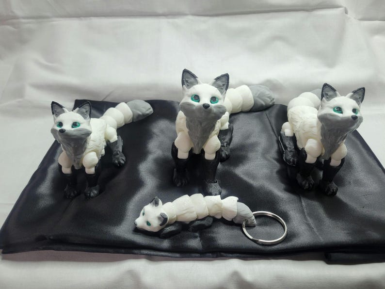 May include: Three white and gray fox figurines with green eyes. The foxes are posed in a sitting position with their tails curled around their bodies. One fox is attached to a silver key chain.