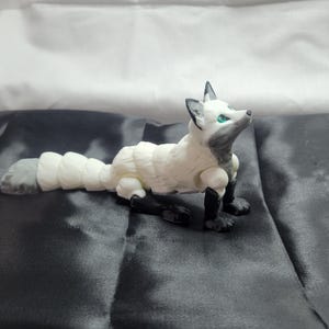 May include: A white and gray articulated fox figurine with blue eyes. The fox is posed lying down with its head tilted up.
