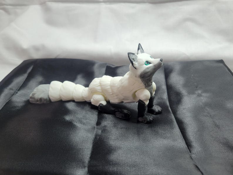 May include: A white and gray articulated fox figurine with blue eyes. The fox is posed lying down with its head tilted up.