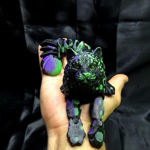 Könnte beinhalten: Eine 3D-gedruckte, bewegliche Leopardenfigur in Schwarz, Grün und Lila. Der Leopard hat grüne Augen und Flecken, mit einem segmentierten Körper und Beinen. Der Schwanz ist lila und grün. Die Figur wird in einer Hand gehalten.
