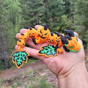 May include: A multi-piece articulated toy featuring a bright orange dragon head with black accents, several small orange jack-o'-lantern beads with black carved faces, and two orange and black maple leaf charms with bright green vein details.