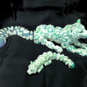 May include: A white sculpture of a cheetah with green and blue spots, lying down with its tail extended. A smaller cheetah sculpture is also present. The sculptures are on a black surface.
