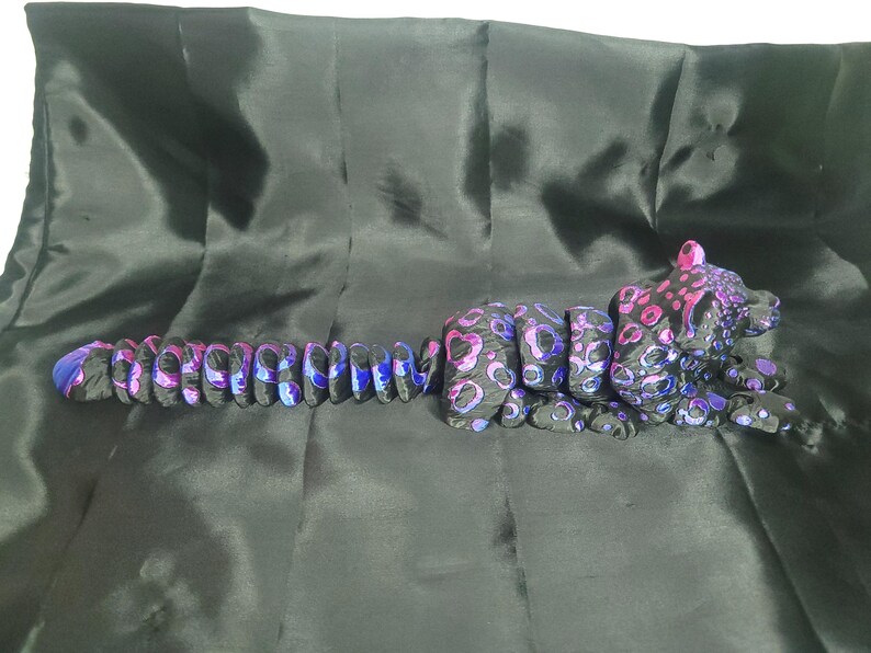 May include: A black and purple leopard sculpture with a segmented body and a spotted pattern. The leopard's body is made up of connected pieces, and the spots are in shades of pink and purple. The sculpture is lying on a black surface.