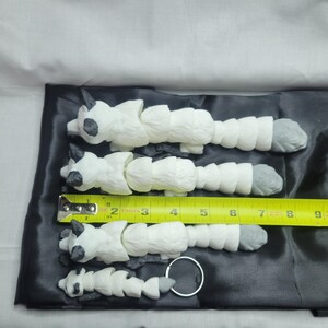 May include: Four white segmented animal figurines with black eyes and gray feet. The figurines are arranged in decreasing size from left to right. A yellow measuring tape is placed below the figurines, with the numbers 2 through 11 visible. The figurines are on a black satin surface.