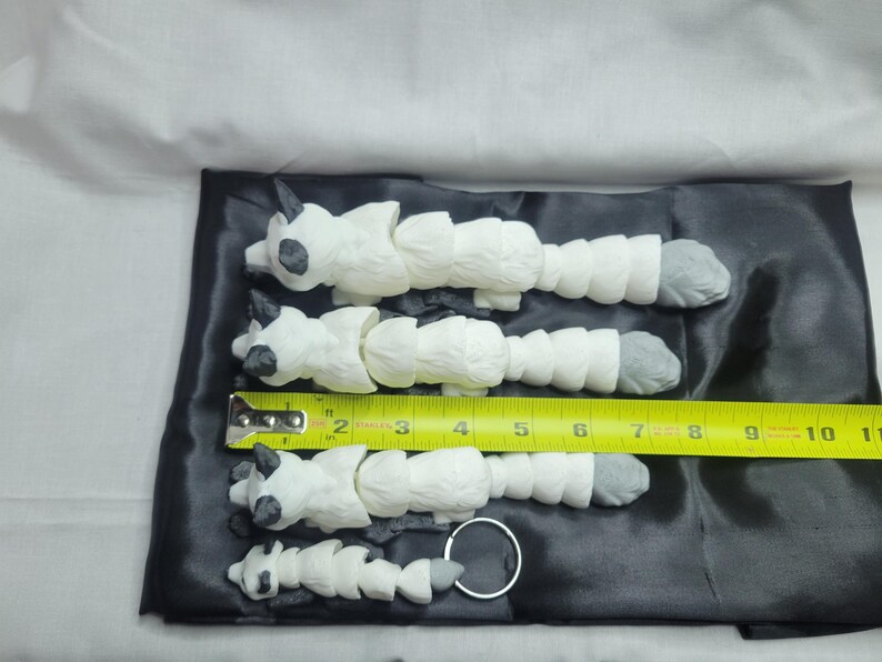 May include: Four white segmented animal figurines with black eyes and gray feet. The figurines are arranged in decreasing size from left to right. A yellow measuring tape is placed below the figurines, with the numbers 2 through 11 visible. The figurines are on a black satin surface.