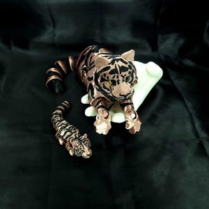 Könnte beinhalten: Zwei Tigerfiguren. Der größere Tiger ist bronzefarben mit schwarzen Streifen und grünen Augen, auf einem weißen Sockel. Eine kleinere Tigerfigur ist ebenfalls bronze mit schwarzen Streifen und grünen Augen. Der Hintergrund ist schwarz.