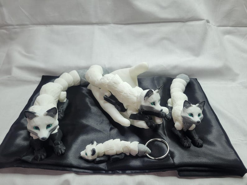 May include: Four white and black fox figurines with green eyes. The foxes are in different poses, including curled up, sitting, and standing. The figurines are on a black fabric background.
