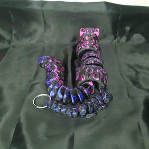 May include: A black and purple beaded keychain with a leopard print design. The keychain is made of multiple connected beads in shades of black, purple, and blue, with a silver key ring.