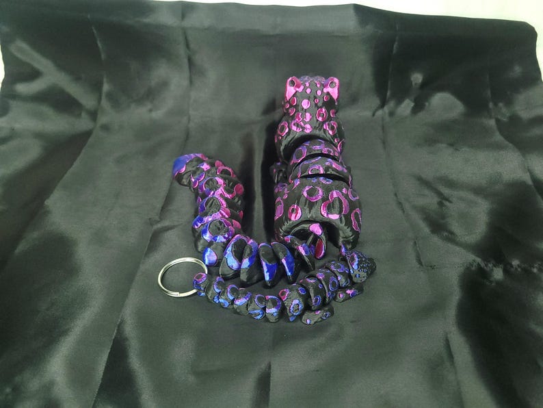 May include: A black and purple beaded keychain with a leopard print design. The keychain is made of multiple connected beads in shades of black, purple, and blue, with a silver key ring.