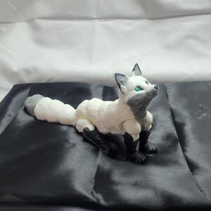 May include: A white and gray articulated fox figurine with green eyes. The fox is lying on a black satin surface.