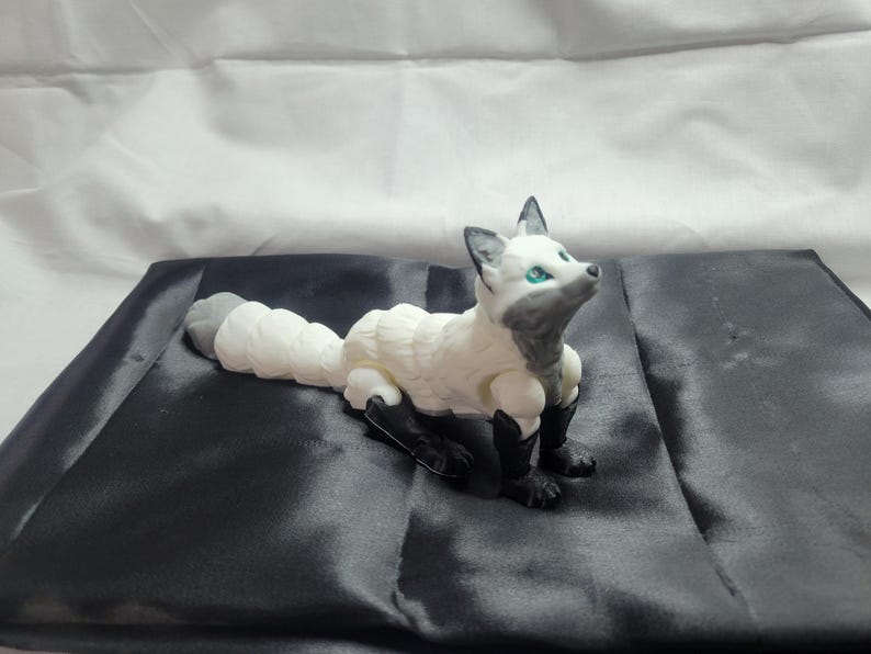 May include: A white and gray articulated fox figurine with green eyes. The fox is lying on a black satin surface.