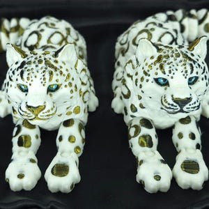 Könnte beinhalten: Zwei weiße und goldene Leoparden-Skulpturen mit blauen Augen werden auf einer schwarzen Oberfläche präsentiert. Die Skulpturen haben ein geflecktes Muster und sind in liegender Position dargestellt. Die Schwänze sind gebogen.