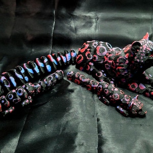Könnte beinhalten: Schwarze Skulptur eines Leoparden mit langem Schwanz, verziert mit rosa und roten Kreisen. Der Schwanz ist mit blauen und rosa Akzenten segmentiert. Die Skulptur liegt auf einer dunklen, strukturierten Oberfläche.