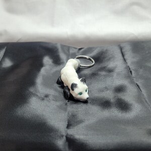 May include: White and black fox keychain with green eyes. The fox is curled up in a playful pose. The keychain is made of plastic and has a silver metal ring.