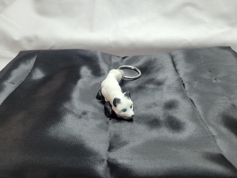 May include: White and black fox keychain with green eyes. The fox is curled up in a playful pose. The keychain is made of plastic and has a silver metal ring.