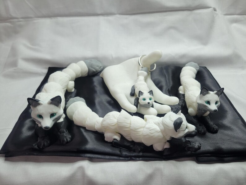 May include: Four white and gray ceramic fox figurines with green eyes. The foxes are arranged on a black satin fabric. One fox is holding a key chain.