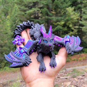 May include: A black articulated dragon figurine with vibrant purple accents on its wings, ears, and tail. The creature features detailed scales, sharp claws, and bright blue eyes, designed with a multi-jointed body structure for movement.
