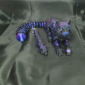 May include: A black and purple articulated jaguar figurine with a matching keychain. The jaguar has a detailed design with purple and blue accents. The tail of the jaguar is a flexible, segmented design. The keychain is a miniature version of the jaguar.