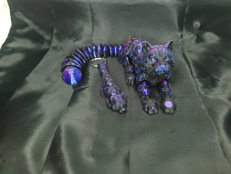 May include: A black and purple articulated jaguar figurine with a matching keychain. The jaguar has a detailed design with purple and blue accents. The tail of the jaguar is a flexible, segmented design. The keychain is a miniature version of the jaguar.