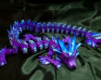 May include: A purple and blue articulated dragon figurine. The dragon has a detailed design with a purple body and blue accents on the head, spine, and feet. The dragon is posed on a dark green fabric.