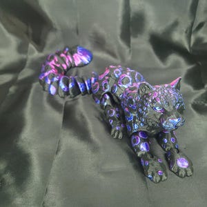 May include: A black and purple jaguar figurine with a long, curved tail. The jaguar has a spotted pattern in shades of purple and blue. The figurine is posed in a crouching position.