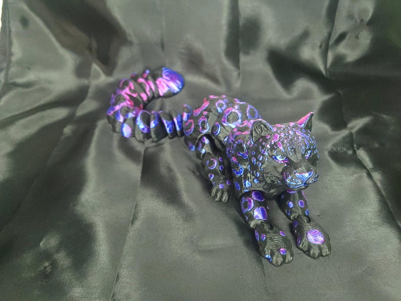 May include: A black and purple jaguar figurine with a long, curved tail. The jaguar has a spotted pattern in shades of purple and blue. The figurine is posed in a crouching position.