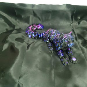May include: A black and purple leopard figurine with a spotted pattern, lying on a dark green satin fabric. The leopard's spots are a mix of purple and blue, and its tail is curved. The figurine has detailed features.