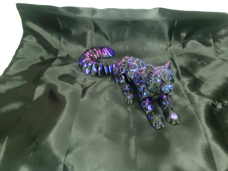 May include: A black and purple leopard figurine with a spotted pattern, lying on a dark green satin fabric. The leopard's spots are a mix of purple and blue, and its tail is curved. The figurine has detailed features.