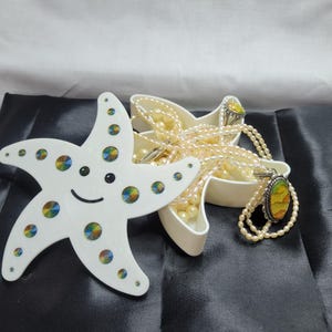 May include: A white starfish-shaped trinket dish with a smiling face and colorful iridescent dots. A second starfish-shaped dish holds a pearl necklace and a pendant necklace with a yellow and orange stone. A ring with a yellow stone is also in the dish.