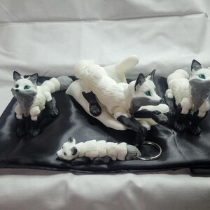May include: Four white and gray fox figurines with green eyes. The foxes are posed in different positions, including sitting, lying down, and standing. The figurines are on a black satin surface.