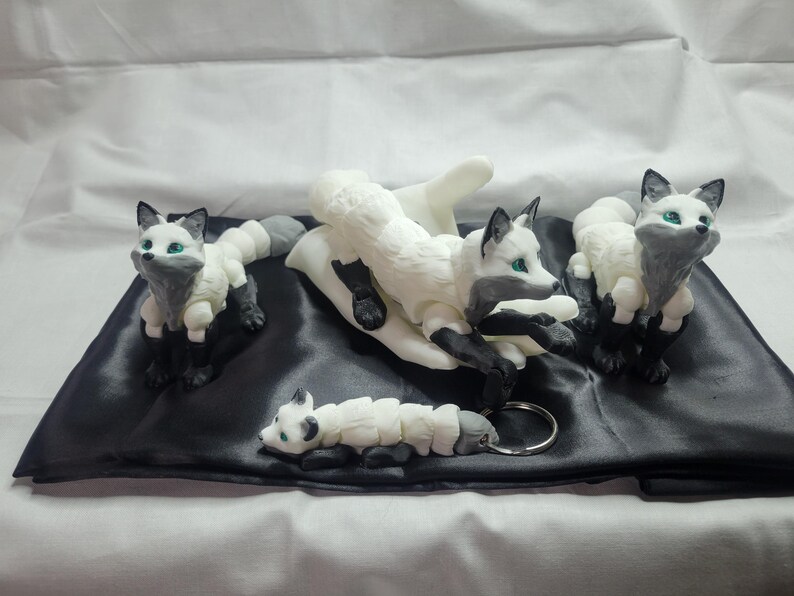 May include: Four white and gray fox figurines with green eyes. The foxes are posed in different positions, including sitting, lying down, and standing. The figurines are on a black satin surface.