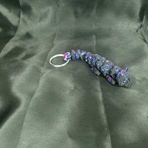 May include: A black and blue jaguar keychain with a silver key ring. The jaguar has blue and purple circular patterns. The keychain is lying on a dark green, silky fabric.