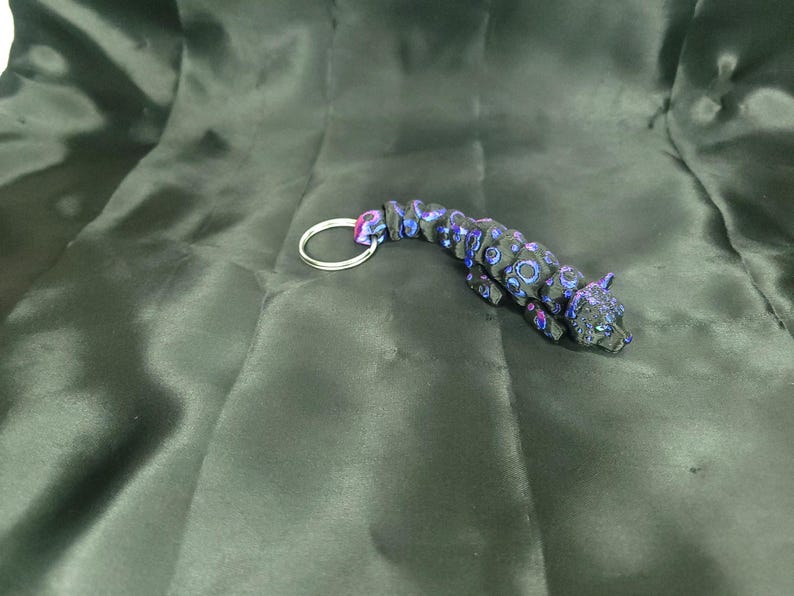 May include: A black and blue jaguar keychain with a silver key ring. The jaguar has blue and purple circular patterns. The keychain is lying on a dark green, silky fabric.