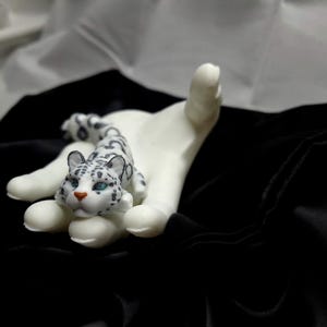 Puede incluir: Una mano de cerámica blanca sosteniendo una pequeña figura de leopardo de las nieves blanca y negra. La figura tiene ojos azules y una cara detallada.
