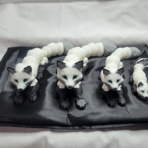 May include: Three white and black fox figurines with green eyes, arranged in a row on a black satin surface. The smallest fox is attached to a silver key chain.