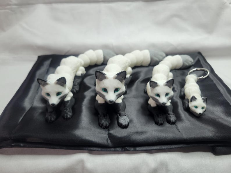 May include: Three white and black fox figurines with green eyes, arranged in a row on a black satin surface. The smallest fox is attached to a silver key chain.