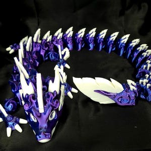 May include: A 3D-printed articulated dragon toy in shades of purple and blue with white accents. The dragon has a detailed design with a long, flexible body and a head with horns and a snout. The toy is on a black surface.