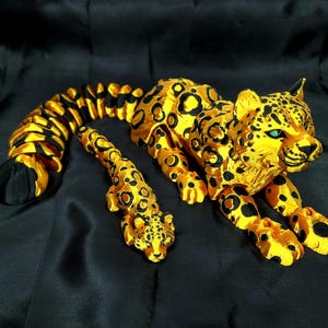 Könnte beinhalten: Goldene Leoparden-Skulptur mit schwarzen Flecken und blauen Augen, auf schwarzem Untergrund. Die Skulptur umfasst ein kleineres Leopardenjunges, beide mit detaillierten Merkmalen und strukturierter Oberfläche.
