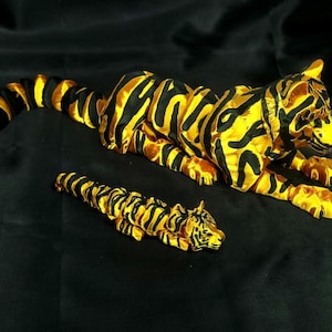 Könnte beinhalten: Zwei dekorative Tigerfiguren. Der größere Tiger liegt, der kleinere darunter. Beide sind gelb mit schwarzen Streifen, vor einem schwarzen Hintergrund.