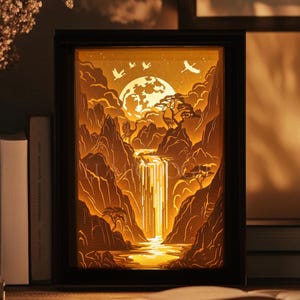 May include: A framed, illuminated landscape art piece featuring a waterfall, mountains, and a full moon. The artwork is in a black frame and sits on a wooden surface with books and a cup. The word "LUMIOLOGIE" is at the bottom.
