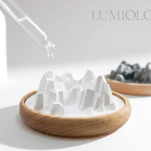 May include: A white ceramic mountain range sits in a wooden bowl, with a dropper dispensing liquid above it. A second bowl with a dark gray mountain range is in the background. The word "LUMIOLOGIE" is in the upper right corner.