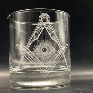 May include: Clear glass tumbler with a detailed etched design. The design features a square and compass symbol with an eye in the center, surrounded by rays. The glass is cylindrical and appears to be empty.