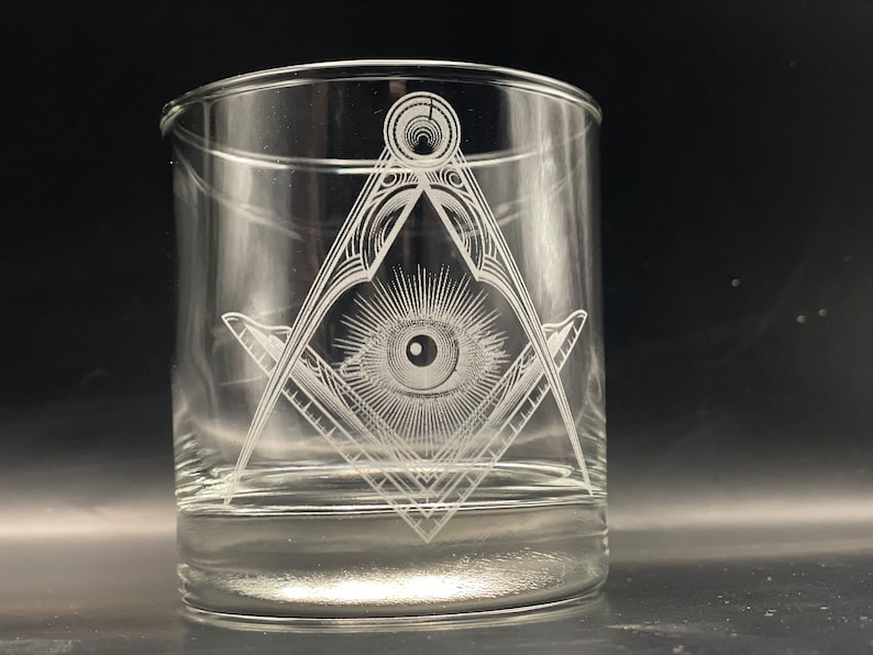 May include: Clear glass tumbler with a detailed etched design. The design features a square and compass symbol with an eye in the center, surrounded by rays. The glass is cylindrical and appears to be empty.