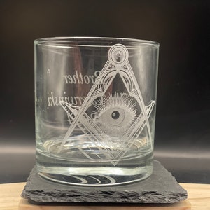 May include: Clear glass tumbler with etched Masonic symbols, including a square and compass with an all-seeing eye. The words "Brother" and "Initiate" are etched on the glass. The glass sits on a dark gray slate coaster.